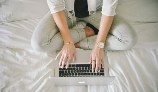 Mujer sentada sobre la cama escribiendo con un portátil.