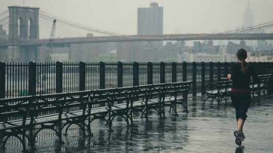 Mujer corriendo junto al río con un puente al fondo en un día lluvioso