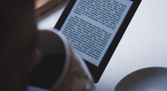 Detalle de un lector con un e-reader leyendo un ebook mientras toma una taza de café.