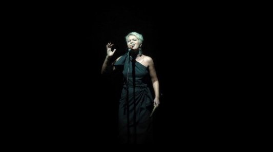 Mujer cantando con un micrófono sobre un escenario negro.