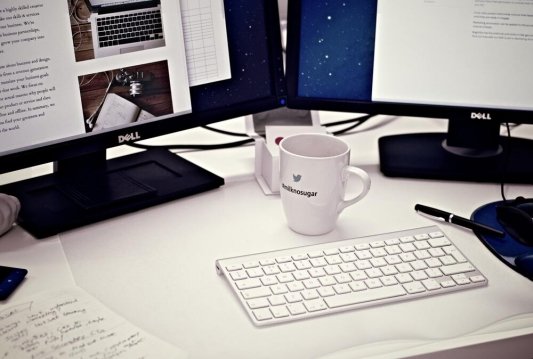 Una mesa de trabajo con una taza, un teclado y dos pantallas