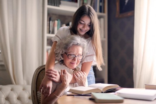 Una nieta abraza a su abuela por la espalda mientras ambas leen un libro abierto sobre la mesa