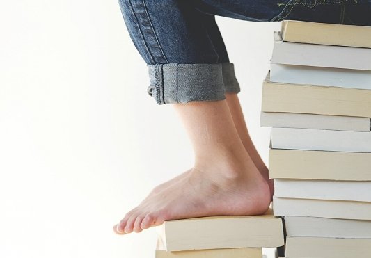 Pila de libros y sobre ella persona a la que solo se le ven las piernas y los pies