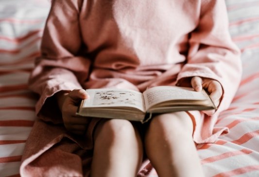 Chica vestida de rosa y sentada sobre una cama sostiene un libro abierto en su regazo