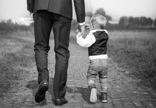 Fotografía en blanco y negro de un padre con traje llevando a su hijo de la mano por un camino