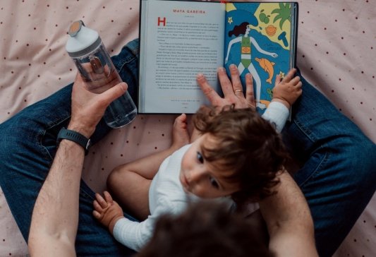 Padre e hijo sentados en una cama mientras el adulto sujeta un libro abierto y el niño le mira