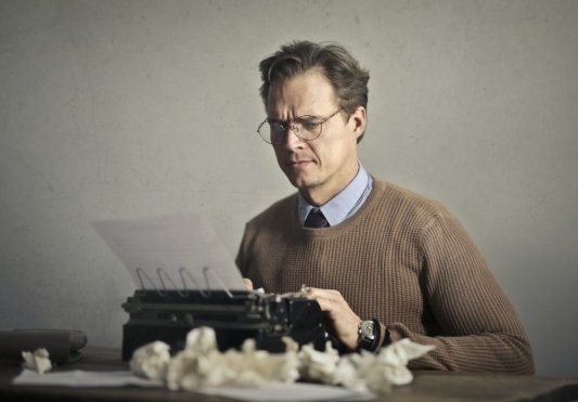 Hombre con gafas y el ceño fruncido escribiendo en una máquina de escribir rodeado de papeles arrugados