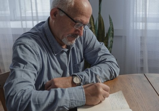 Hombre mayor escribe en un cuaderno apoyándose en una mesa
