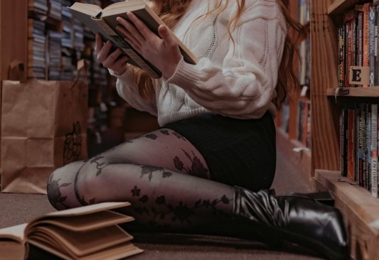 Chica sentada en el suelo de una biblioteca con un libro abierto frente a ella y otro que está leyendo en las manos