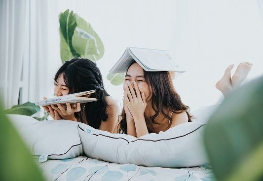 Dos chicas tumbadas sobre una cama riendo mientras una sostiene un libro en las manos y la otra otro abierto sobre su cabeza