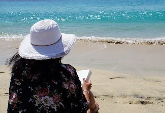 Fotografía de una mujer de espaldas con sombrero sentada en la arena frente al mar leyendo un libro