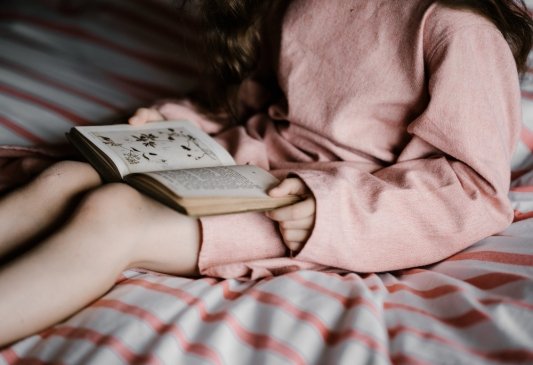 Niña con un vestido rosa sentada sobre un acama leyendo un libro abierto apoyado sobre su regazo