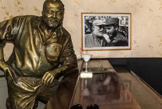 Escultura de Ernest Hemingway en una barra de bar acompañado de una copa, detrás una foto en blanco y negro del autor